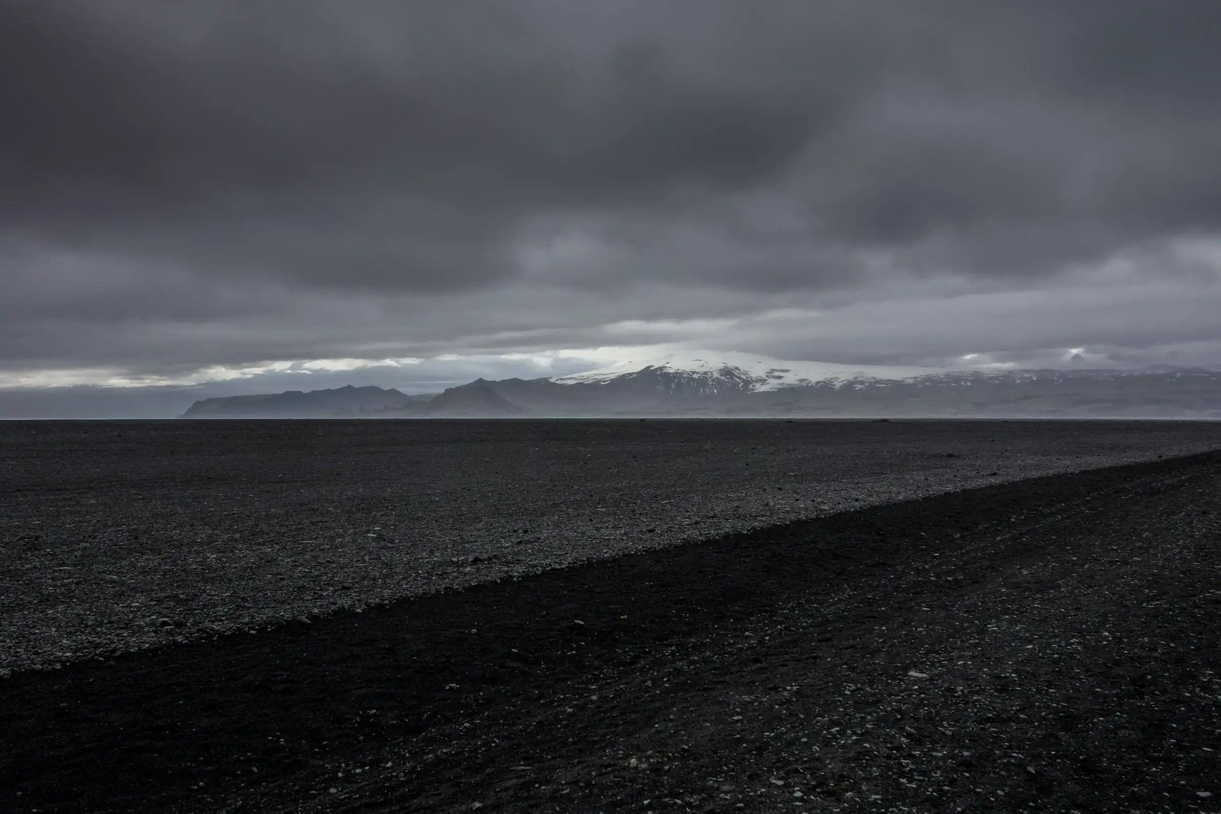 A dark gravel plain under heavy cloud, with distant mountains on the horizon