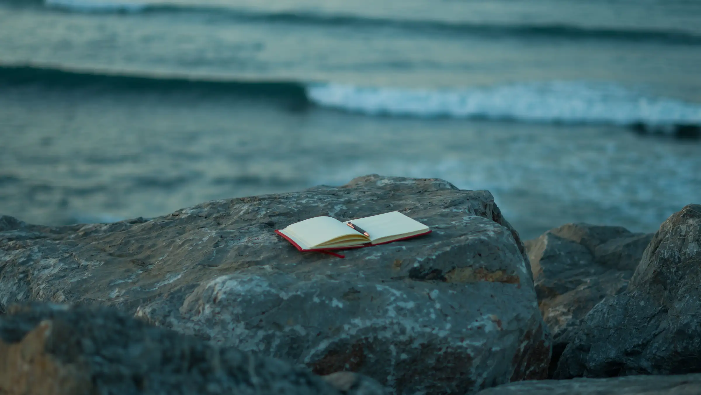 An open notebook and pen resting on a rock above a grey sea at dusk